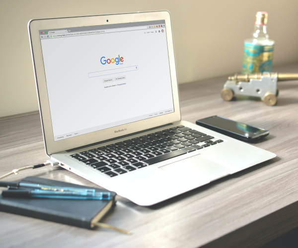 A MacBook Air laptop displaying the Google search engine on its screen is open on a light wooden desk, with a smartphone, a notebook with a pen, a decorative cannon, and a bottle behind it.