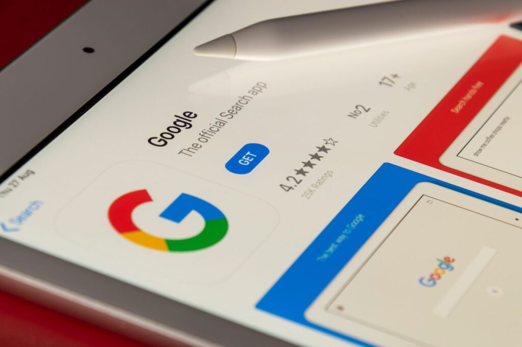 Tablet screen displaying the Google app icon and information, with a stylus resting on the screen. The app listing shows a 4.2-star rating, along with "GET" button. Other app features are visible on the screen. The background is red.