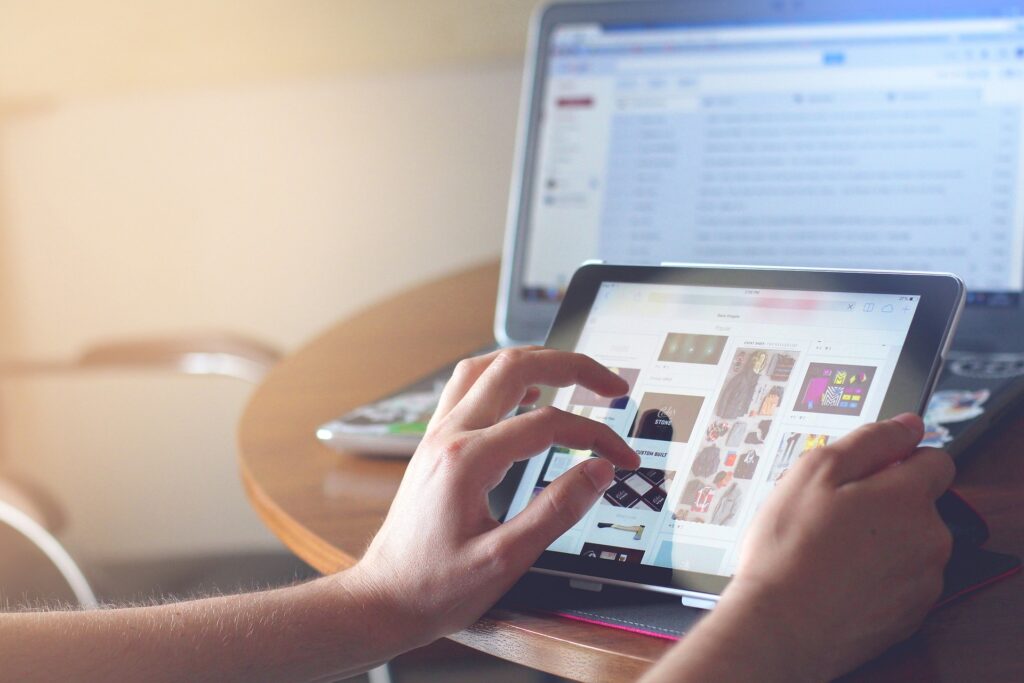 Hands using a digital tablet displaying website design ideas, with a laptop computer in the background, all on a wooden table, suggesting digital marketing and website development work.