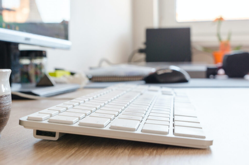 White computer keyboard on a wooden desk with a computer monitor, laptop, mouse, and other office supplies in the background.
