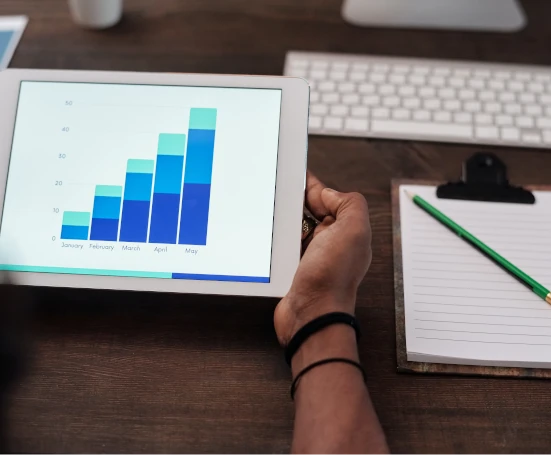 Person holding a white tablet displaying a stacked bar graph on a wooden desk. The graph shows a positive trend from January to May. A keyboard, notepad with a green pencil, and a coffee cup are visible in the background.