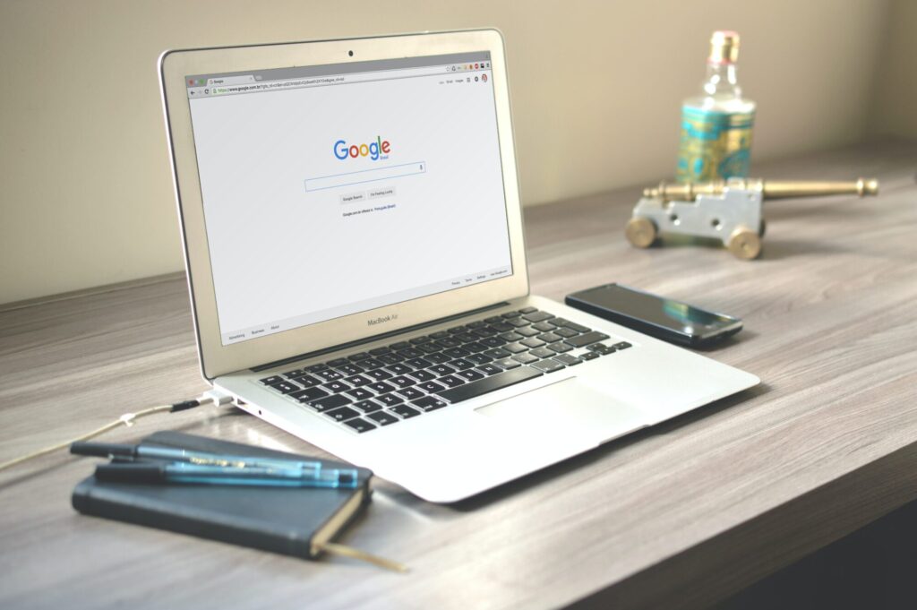 Laptop displaying the Google search page open on a wooden desk, along with a smartphone, notebook, pens, and decorative objects.