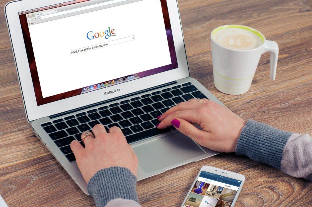 A person typing on a silver MacBook Air laptop with the Google search engine open, searching for "Wilof, Free photo, mockups, crm". A smartphone displaying photos is to the right of the laptop. A white mug with coffee sits to the right of the laptop, on a wooden surface. The person's hands are visible on the keyboard. The person is wearing a gray sweater.