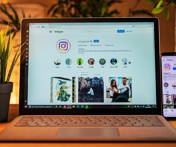 Laptop and smartphone displaying the Instagram interface. The laptop screen shows the Instagram logo, profile information, and a selection of user posts. A smartphone to the right of the laptop also shows the Instagram interface. A potted plant is to the left of the laptop. The devices are positioned on a wooden surface, suggesting a workspace.