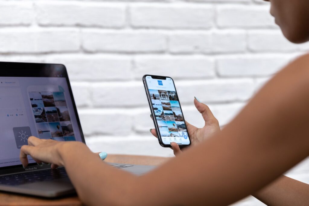 A person using a Macbook Pro laptop and a smartphone against a white brick wall, reviewing images for social media or website content, possibly digital marketing or website development.