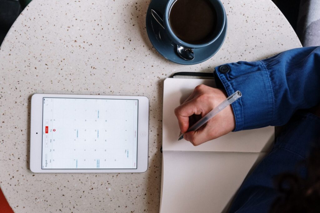 Person writing in a notebook next to a tablet displaying a calendar, with a cup of coffee on a speckled table. The tablet displays the calendar for August 2020. The person's hand, wearing a blue sleeve, holds a pen.