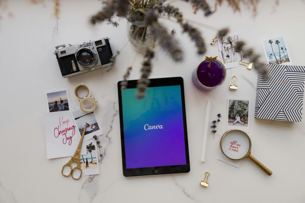 An overhead shot of a marble surface with a tablet displaying the Canva logo, a vintage camera, postcards, a notebook, gold paper clips, scissors, and a magnifying glass. Other elements include washi tape, a candle, a white stylus, and a small vase of dried lavender.