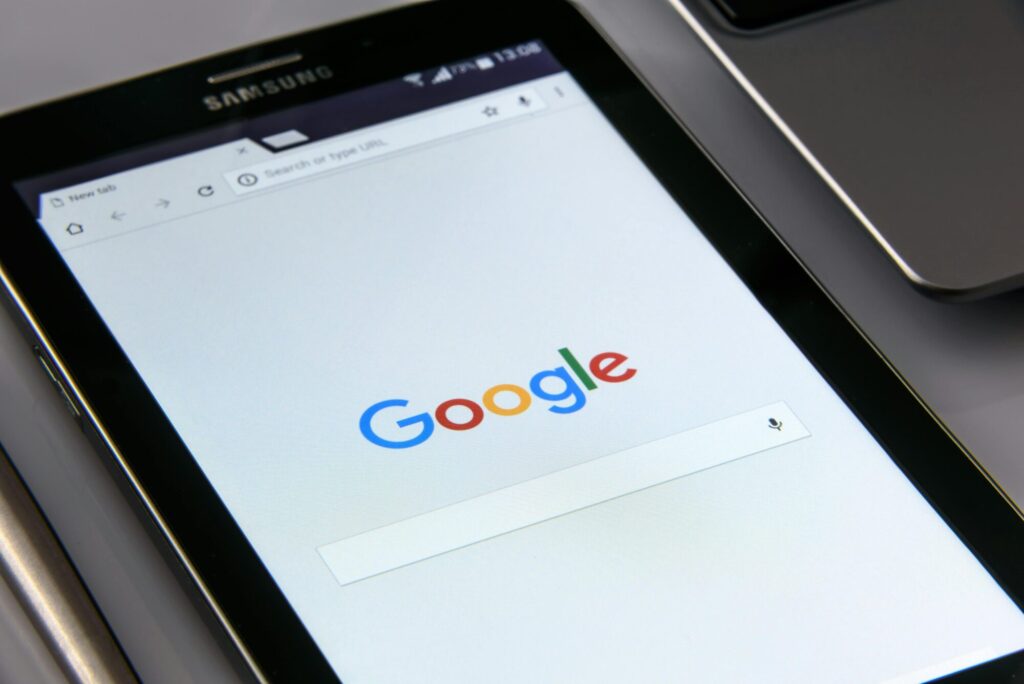 A black Samsung tablet displaying the Google search page on its screen, with part of a laptop visible in the background. The tablet screen shows the Google logo and a search bar.