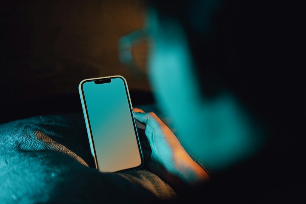 A person holding a smartphone with a blank screen, lit by blue and orange light, set against a dark background. The hand and phone are in focus, with the screen reflecting the cool tones. A patterned fabric is visible beneath the phone.