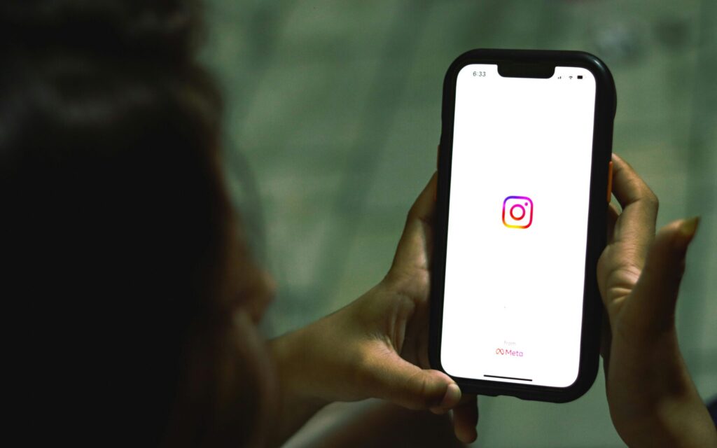 A person holding a smartphone displaying the Instagram app logo on the screen. The logo is the classic colorful gradient square with a rounded-corner camera icon in the center. The phone is in focus, while the person's face and the background are blurred. The time 6:33 is visible at the top of the screen. The Meta logo is shown under the Instagram logo.
