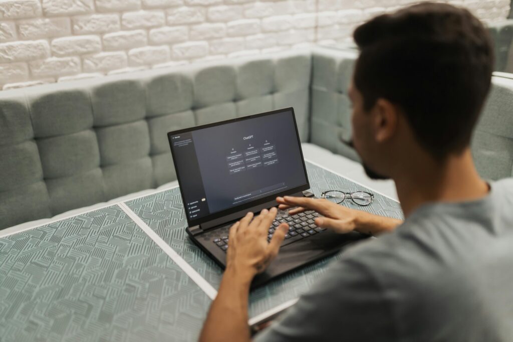 Person working on a laptop displaying ChatGPT interface, typing on keyboard. The person is seated at a table with a patterned surface, with eyeglasses and a grey wall behind them. A padded bench is visible behind the person and table.