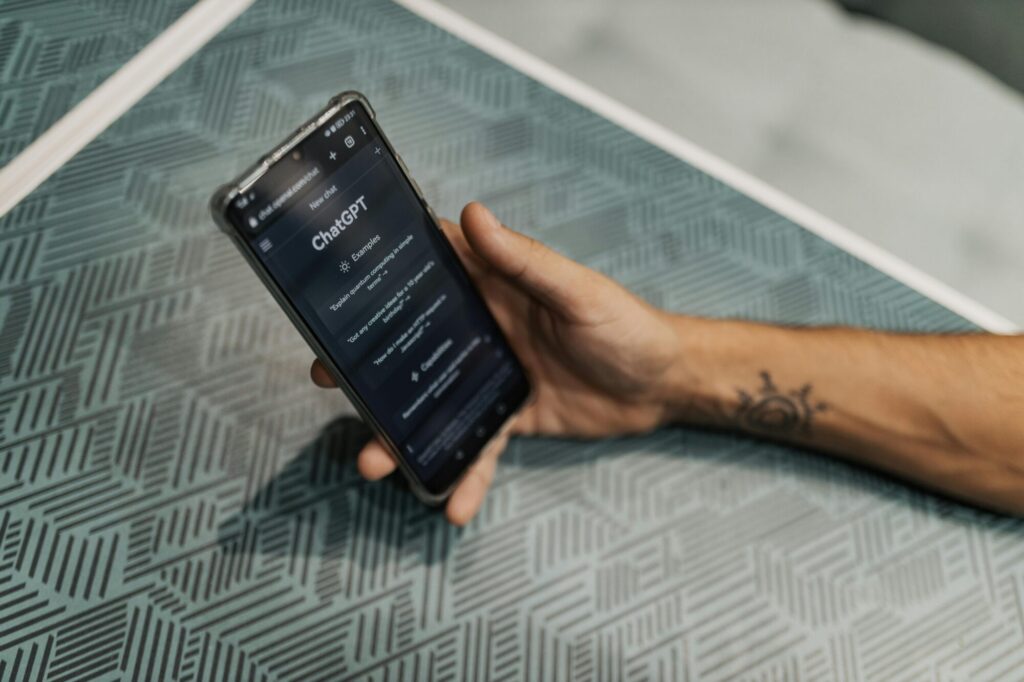 A hand holding a smartphone displaying the ChatGPT interface on a patterned surface. The phone's screen shows the ChatGPT logo and example prompts like "Explain quantum computing in simple terms". A small tattoo is visible on the wrist.