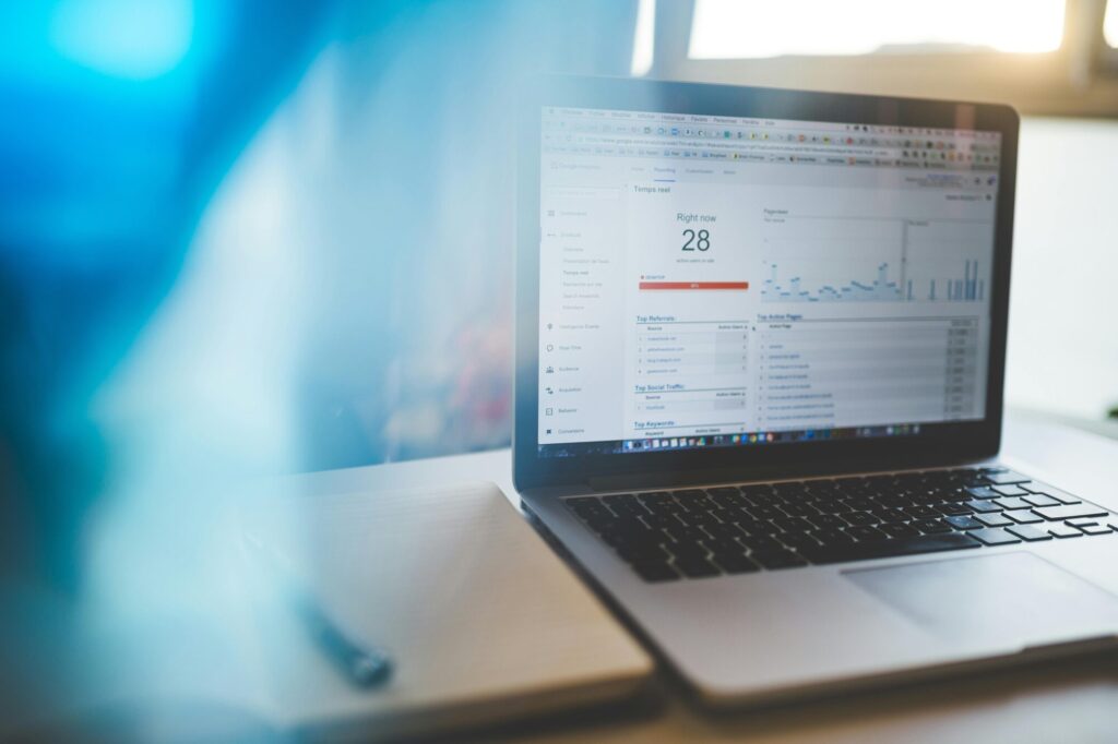 Laptop displaying a Google Analytics dashboard with website traffic data, alongside a notepad and pen. The screen shows real-time website visitors and referral information, likely representing digital marketing analysis and website development activities.