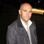 Bald man wearing a grey jacket over a white collared shirt and grey sweater, slightly smiling, with a dark background and a blurred car in the distance.