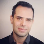 Headshot of a man with dark hair, brown eyes, and a slight smile, wearing a black jacket over a dark gray shirt, against a light background.