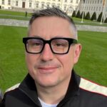 Headshot of a man with short graying hair wearing black square glasses, a black jacket, standing in front of a building with manicured lawn and trees.