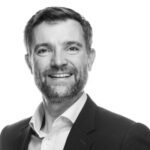Black and white headshot of a smiling man with short hair and a neatly trimmed beard, wearing a suit jacket and collared shirt, likely a professional in digital marketing or web development.