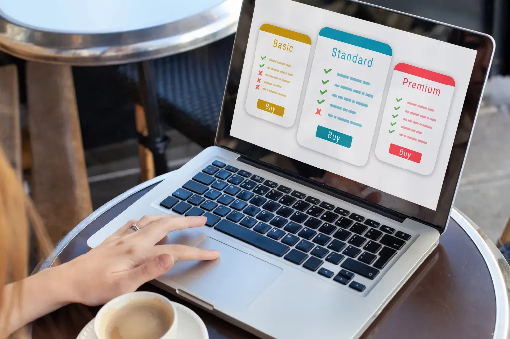 Person using a laptop displaying a pricing comparison chart for digital marketing or website development services. The laptop screen shows three plan options: Basic, Standard, and Premium, each with different features represented by checkmarks and X marks. The plans include "Buy" buttons. A hand rests on the laptop touchpad, and a cup of coffee sits beside the laptop on a cafe table.