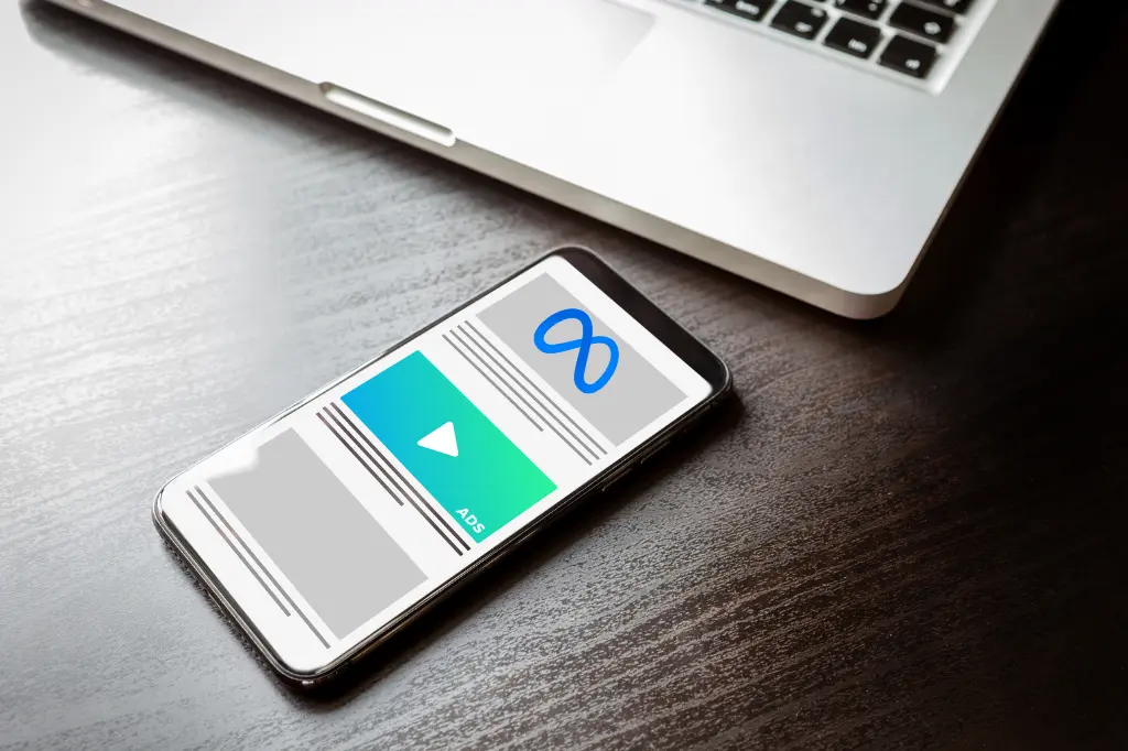 Smartphone displaying a Meta ad, with the Meta logo and a play button, next to a laptop on a dark wooden surface.