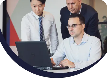 Three business professionals collaborating over a laptop. One man in a light blue shirt is sitting and using the laptop, two other men in shirts and a blazer are looking at the screen with him.
