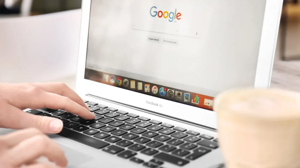 Hands typing on a MacBook Air keyboard with the Google search engine displayed on the screen, a coffee cup is slightly out of focus in the foreground.