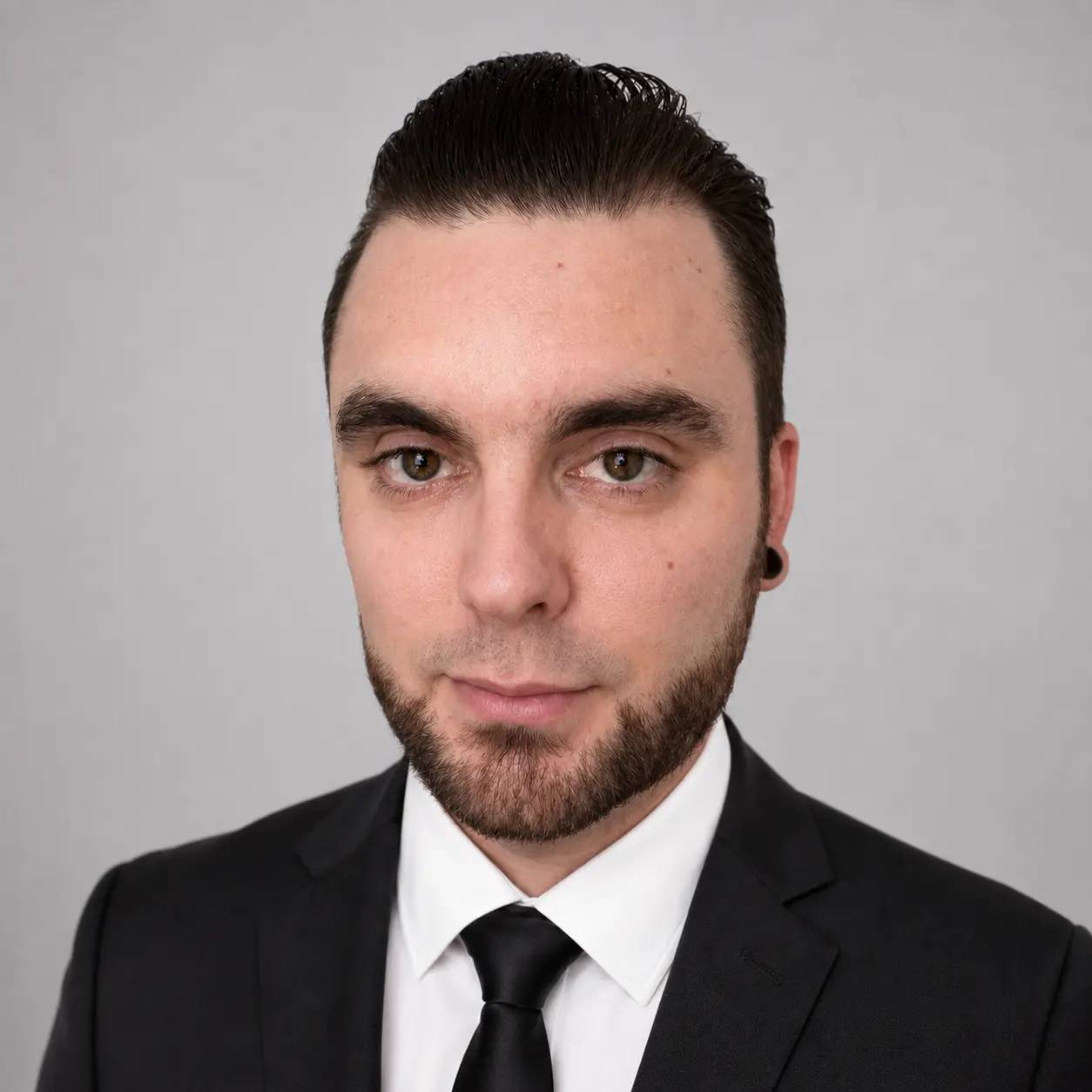 Matt Case with dark hair styled back, wearing a suit and tie, and a small goatee, with a black ear gauge. He is facing the camera.
